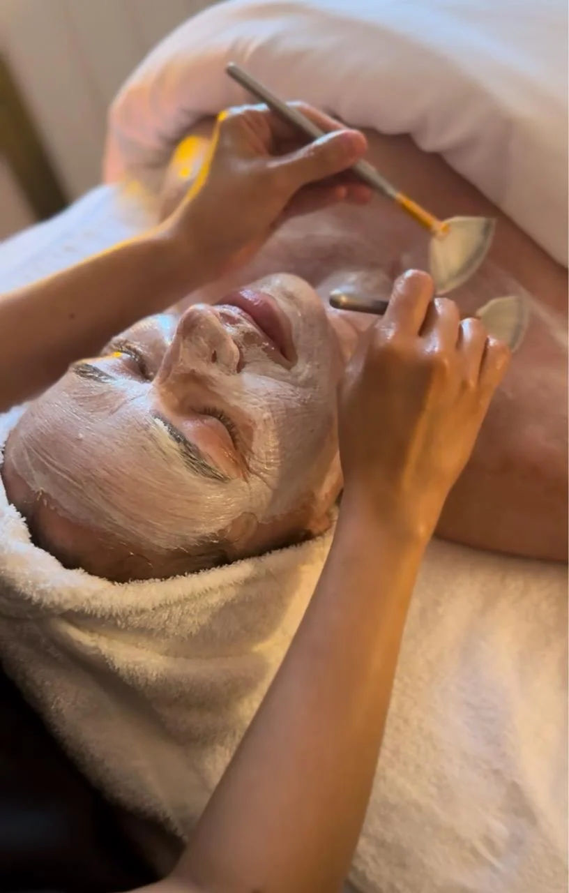 An esthetician brushes a white mask onto a smiling client's face and neck with two fan brushes