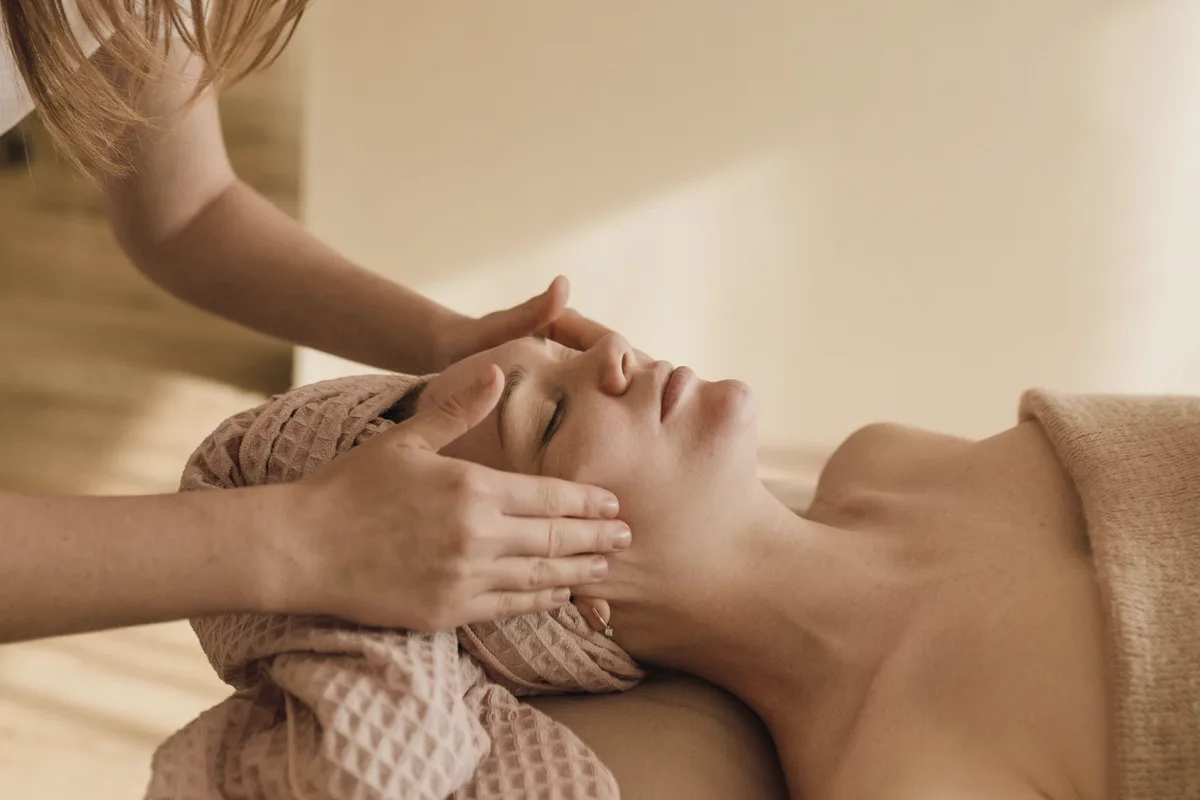 An esthetician rests her fingertips on the temples of a relaxed client wrapped in a waffle-knit towel